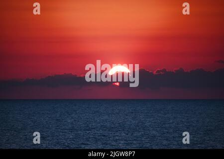 Dramatic sunset over the ocean horizon at dusk with clouds partly covering the sun against a deep red sky background - dreamy glowing light fading int Stock Photo