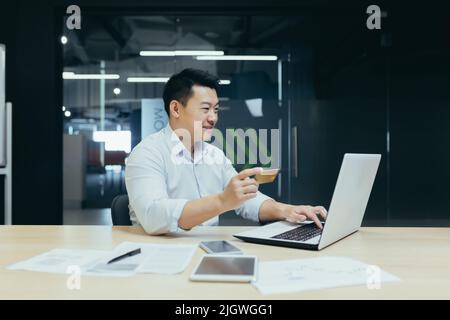 Cheerful and smiling asian businessman working in modern office, dark colors, making online purchase, using bank credit card and laptop, for online shopping Stock Photo