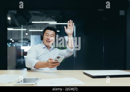 Businessman in office resting during work break, listening to music in big black headphones, Asian man using music app on phone Stock Photo