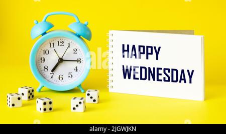 On a yellow background, a blue alarm clock, dice and a notebook with the inscription HAPPY WEDNESDAY. Stock Photo