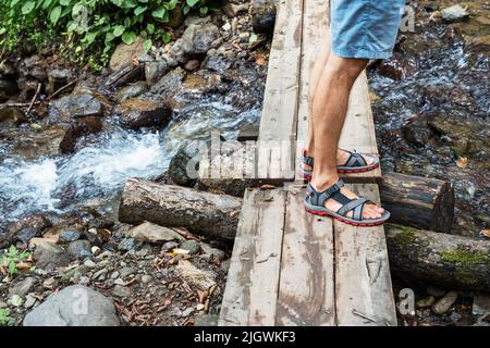male feet in sandals on a wooden bridge across a mountain river man hiking Stock Photo