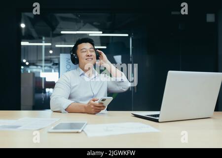 Businessman in office resting during work break, listening to music in big black headphones, Asian man using music app on phone Stock Photo