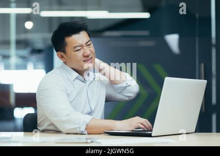 Overtired asian worker in office, having severe neck pain, massaging with hand, businessman working in office with laptop Stock Photo