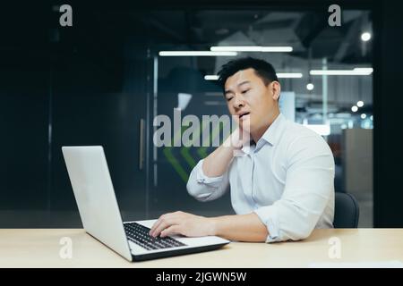 Overtired asian worker in office, having severe neck pain, massaging with hand, businessman working in office with laptop Stock Photo