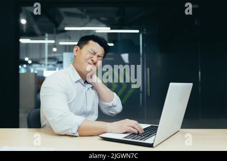 Overtired asian worker in office, having severe neck pain, massaging with hand, businessman working in office with laptop Stock Photo