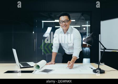 Portrait of successful and experienced Asian architect, man standing over table with plan drawing, smiling and looking at camera, working in modern design studio Stock Photo