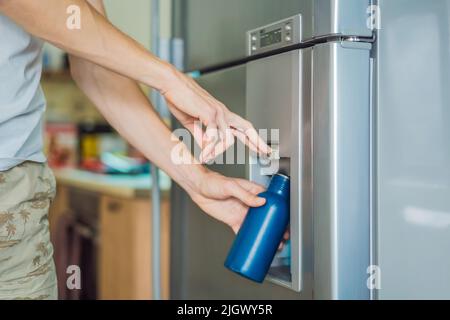 Male hand is pouring cold water and ice cubes in a metal bottle from dispenser of home fridge Stock Photo