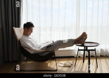 smiling Asian young man reading book on chair near window under sunshine Stock Photo
