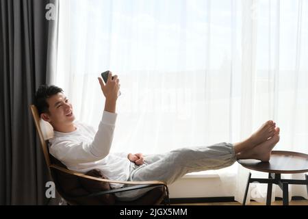 smiling Asian young man sitting on chair near window, using phone to video call Stock Photo