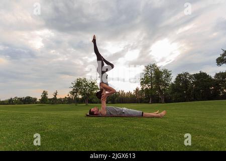 Healthy man lying on grass and balancing woman. Couple doing acrobatic yoga exercise in park Stock Photo