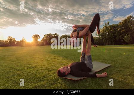 Man lying on grass and balancing woman in his feet. Young couple doing acro yoga in park at sunset Stock Photo