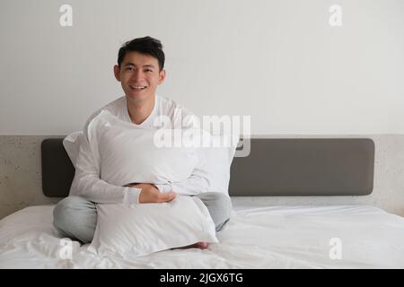 Smiling Asian young man holding a pillow sitting on bed, looking at camera Stock Photo