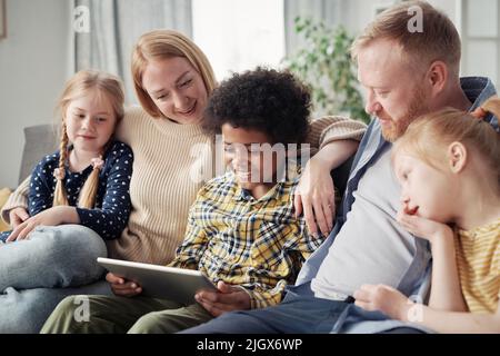 Happy adopted children watching video on digital tablet together with their foster parents during leisure time on sofa at home Stock Photo