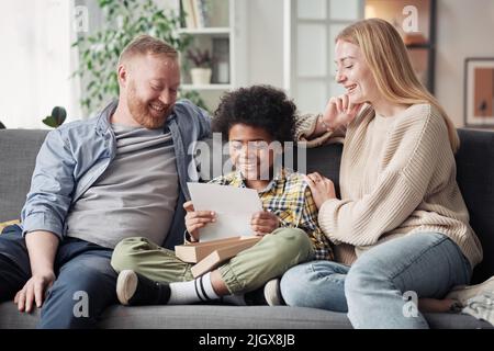 Happy child reading a letter about adoption while sitting on sofa with his foster parents Stock Photo