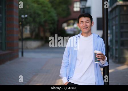 smiling Asian young man holding a cup of coffee on the street, looking at camera Stock Photo
