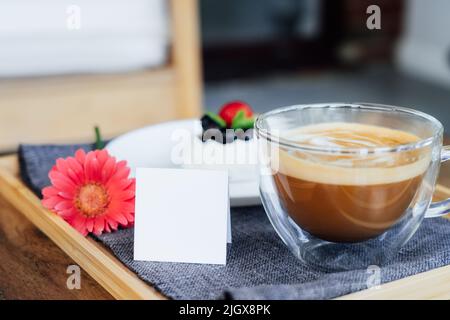 Cup of coffee, Pavlova dessert cake, gerbera flower and blank postcard with copy space on the tray with gray napkin. Breakfast for loved one. Romantic Stock Photo