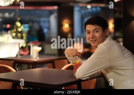 one smiling Asian young man drinking cocktail at bar table, turning around and looking at camera at night Stock Photo
