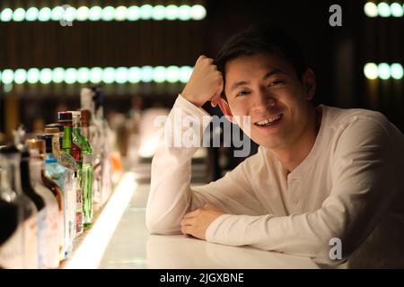 portrait of one smiling Asian young man sitting at bar counter, looking at camera at night Stock Photo