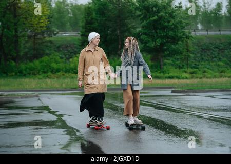 Happy mother and daughter riding on skateboards on asphalt on a rainy day. Holding hands, looking at each other. Green vegetation in background. Stock Photo