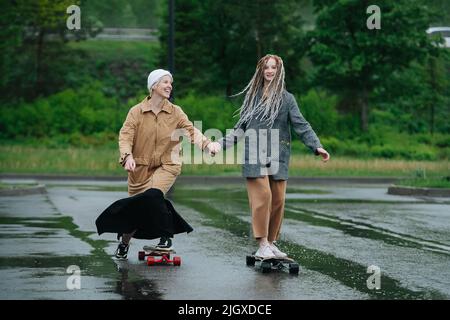 Mother and daughter riding on skateboards on asphalt on a rainy day. Holding hands. Green vegetation in background. Stock Photo