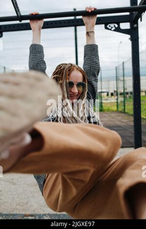 Childish teenage girl with dreads hanging on a high bar, kicking her boot the camera. Smiling, wearing sunglasses. Stock Photo