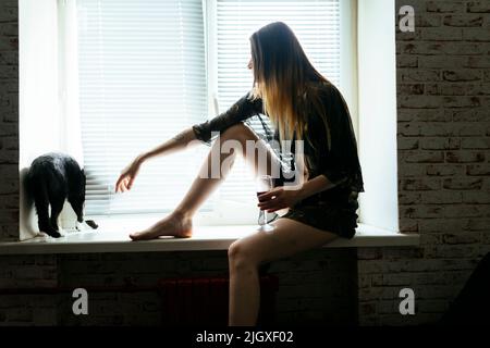 Woman in nightgown sitting on windowsill with glass of wine Stock Photo