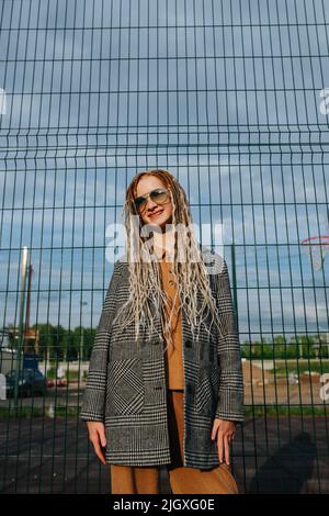 Sunset portrait of a teenage girl with dreads in sunglasses standing in front of a metal fence. She is wearing a grey checkered jacket. Portrait. Stock Photo