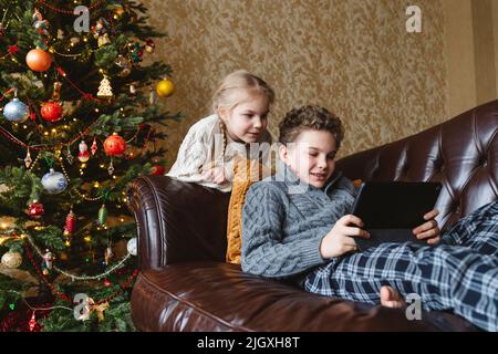 Brother and sister are looking at a tablet near the Christmas tree. Stock Photo