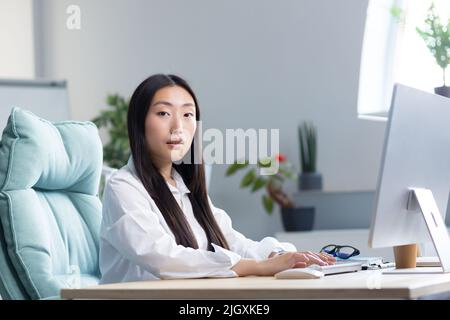Close-up photo. Portrait of a young beautiful Asian woman secretary sitting at the desk at the computer in the office in business clothes, looking at the camera. Stock Photo