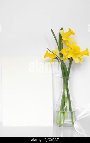 Bouquet of yellow daffodils in a transparent glass vase and an empty white sheet of paper, mock-up Stock Photo