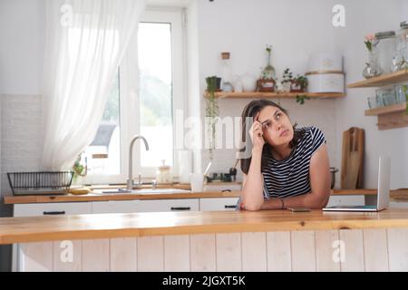 A young woman is in the kitchen in her house. The woman looks thoughtfully to the side, determining what she needs to buy in the store, there is a laptop next to her. High quality photo Stock Photo