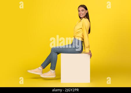 Woman Smiling To Camera Sitting On Cube Over Yellow Background Stock Photo