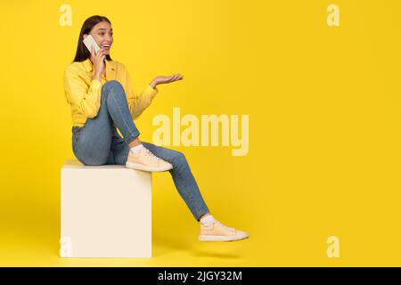 Woman Talking On Phone Sitting On Cube Over Yellow Background Stock Photo
