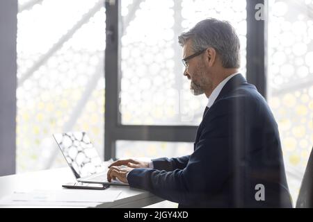 Online Correspondence. Smiling Mature Male Entrepreneur Working With Laptop In Modern Office Stock Photo