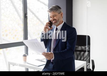 Smiling Middle Aged Businessman Talking On Cellphone And Checking Papers In Office Stock Photo