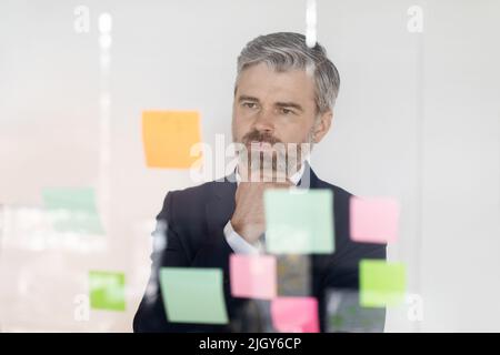 Pensive Businessman Looking At Sticky Paper Notes On Glass Wall In Office Stock Photo