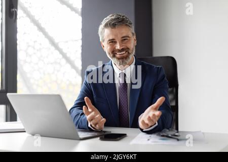 Successful Middle Aged Businessman Sitting In Office, Smiling And Gesturing At Camera Stock Photo
