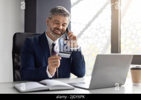 Middle Aged Businessman Talking On Cellphone And Using Credit Card In Office Stock Photo