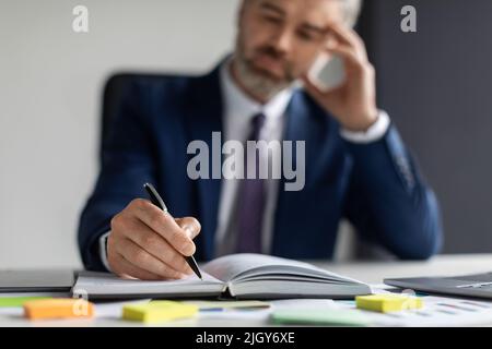 Middle Aged Businessman Writing In Notepad While Sitting At Desk In Office Stock Photo