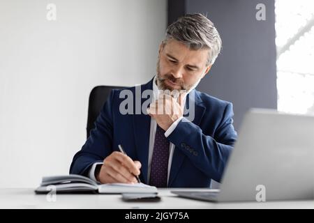 Busy Handsome Middle Aged Businessman Writing In Notepad While Sitting In Office Stock Photo