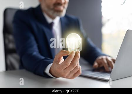 Business Innovations. Businessman Holding Illuminated Light Bulb While Sitting At Workplace Stock Photo