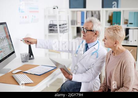 Confident senior male practitioner with gray hair sitting at table and pointing at computer screen while consulting mature female patient in clinic Stock Photo