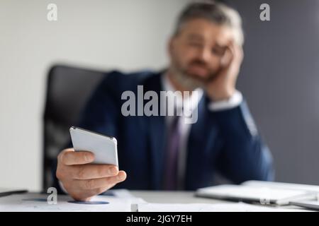 Bored Middle Aged Businessman Sitting At Desk And Holding Smartphone In Hand Stock Photo