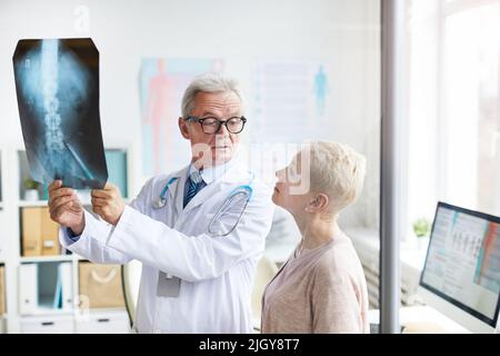 Frowning handsome aged doctor in eyeglasses holding x-ray image against light while explaining spinal problems to senior female patient Stock Photo