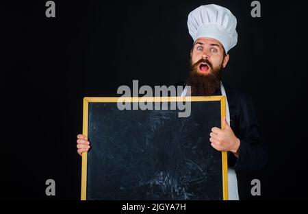 Surprised chef, cook or baker in uniform with empty menu chalkboard show thumb up. Cooking and food. Stock Photo