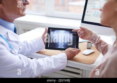 Close-up of female patient sitting table and pointing at tablet screen while asking question about online spine x-ray image to doctor Stock Photo