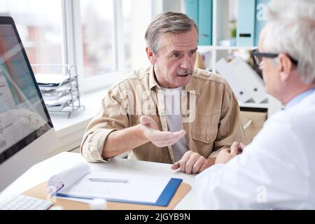 Senior male patient in casual shirt sitting in front of doctor and gesturing hand while discussing medicine with doctor in clinic room Stock Photo