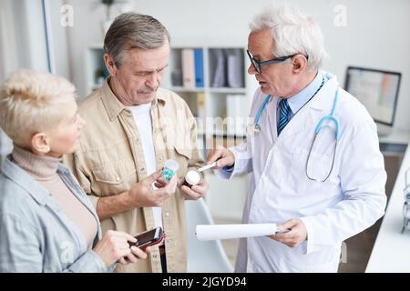 Serious aged male doctor in glasses standing in clinic and explaining senior couple how to take medicine correctly Stock Photo