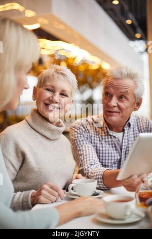 Group of happy senior friends sitting at table and drinking tea while discussing internet news together in cafe Stock Photo