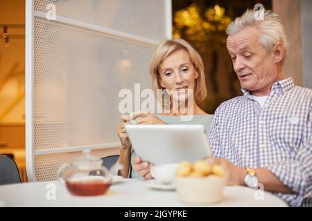 Positive beautiful senior couple sitting at table with tea and sweets and using modern device to watch video Stock Photo